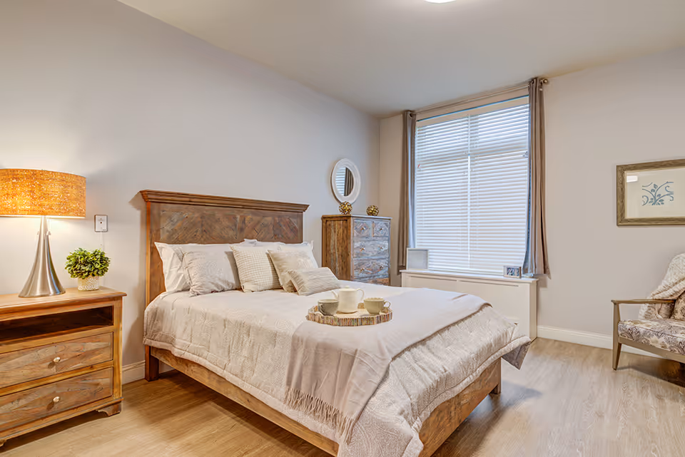 A cozy bedroom with a wooden bed frame, neatly made bed with multiple pillows, a wooden nightstand with a lamp and a small plant, a wooden dresser with decorative items, a large window with blinds and curtains, and a cushioned chair with a throw blanket.