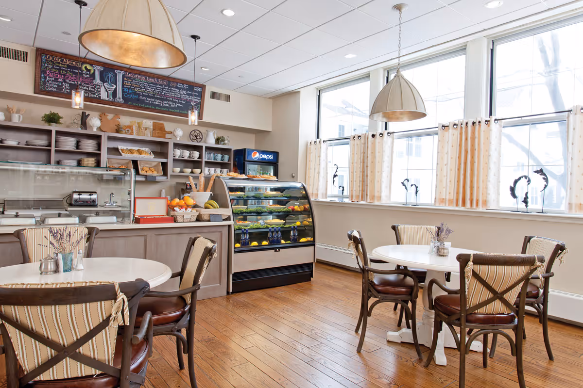 Bright dining area with round tables and chairs, a service counter and refrigerated display case beneath large windows.