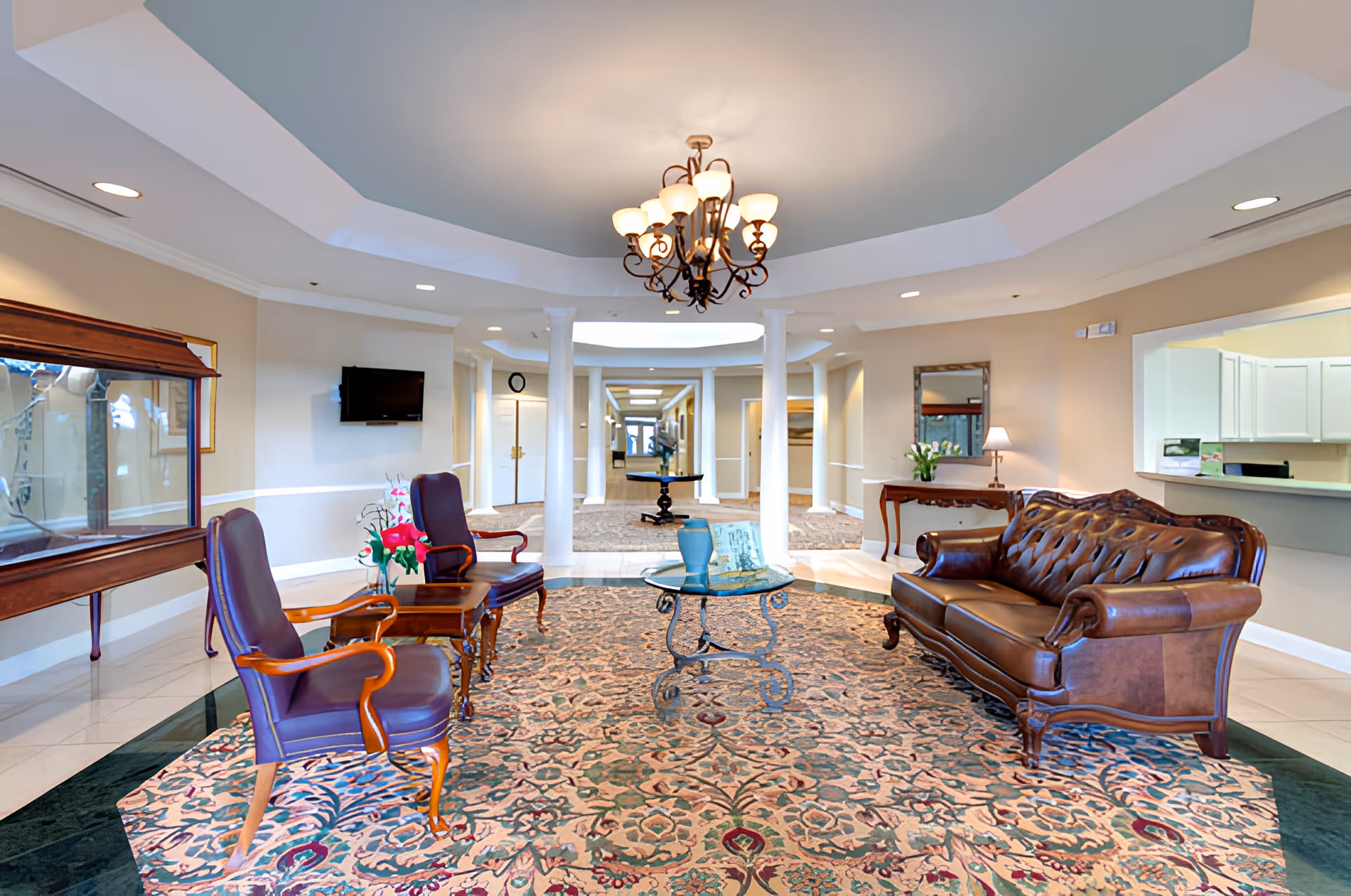 Bright, elegant lobby with a leather sofa, armchairs, glass coffee table on a patterned rug beneath a chandelier.