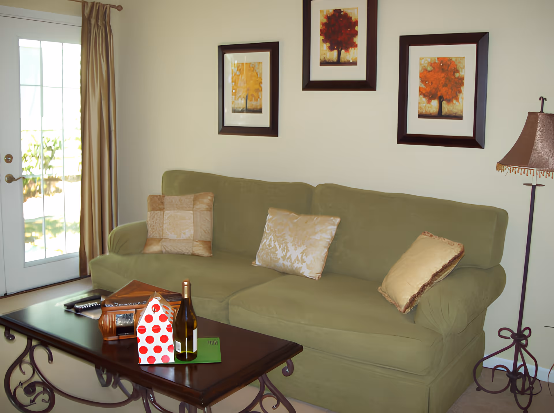 A cozy living room with a green sofa adorned with three decorative pillows. Above the sofa, there are three framed pictures of trees with autumn-colored leaves. To the right of the sofa is a floor lamp with a brown lampshade. In front of the sofa is a wooden coffee table with a bottle of wine, a red and white polka dot gift bag, and some other small items. A glass door with beige curtains is on the left side, letting in natural light.