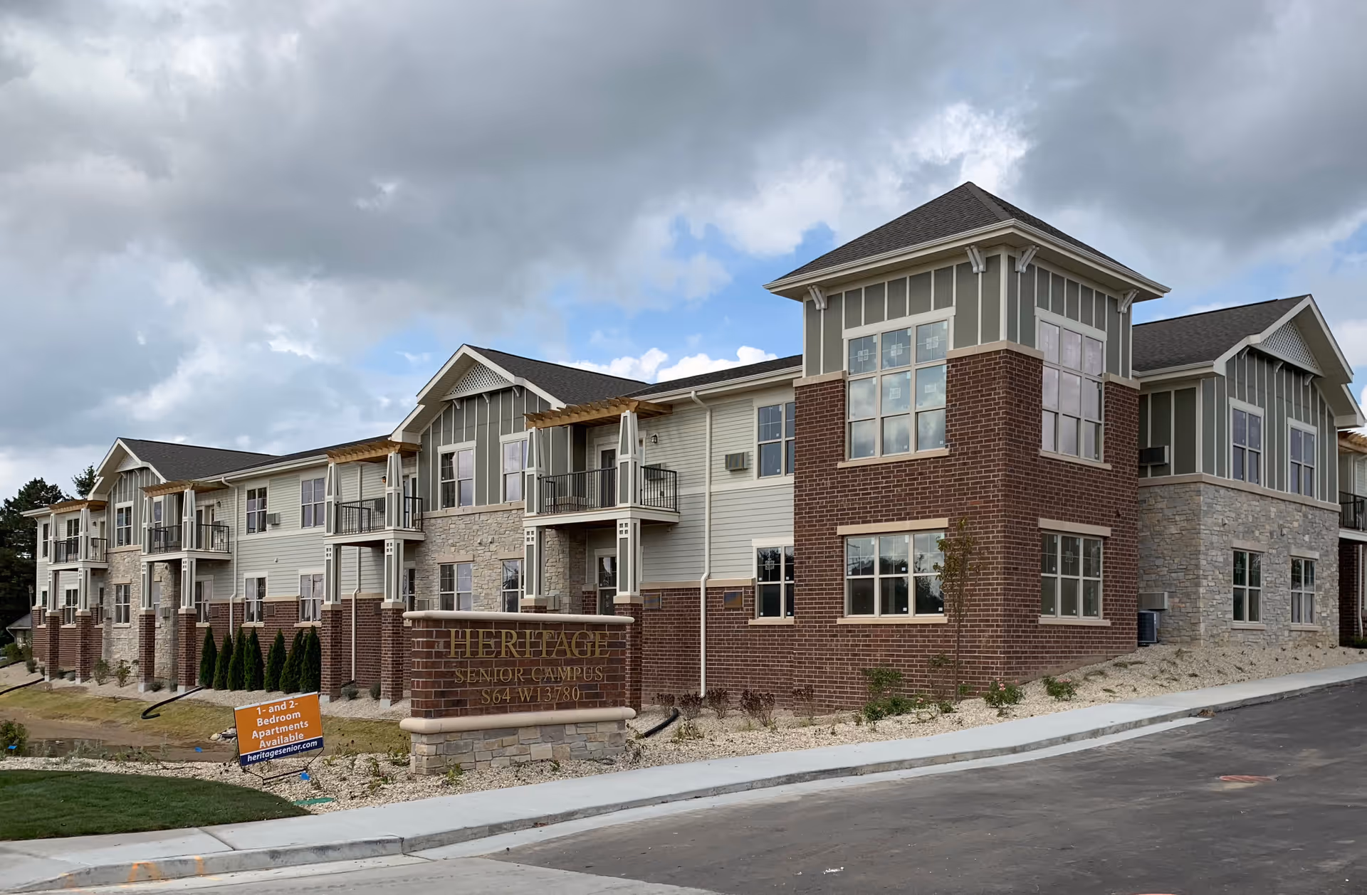 Exterior view of a multi-story senior living facility building with brick, stone, and siding facade under a cloudy sky. There is a sign in front that reads 'HERITAGE SENIOR CAMPUS S64 W13780' and another smaller sign advertising 1- and 2-bedroom apartments available.