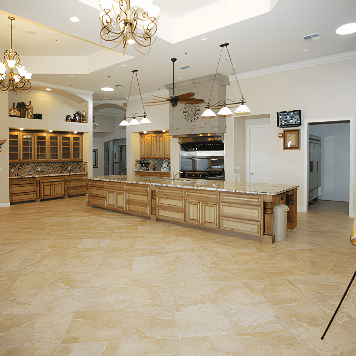 Spacious kitchen area with a large marble countertop island, wooden cabinets, and multiple hanging light fixtures. The floor is tiled with beige stone, and there are decorative elements on the walls and ceiling. The kitchen includes a stove with a large vent hood and a ceiling fan above the island.