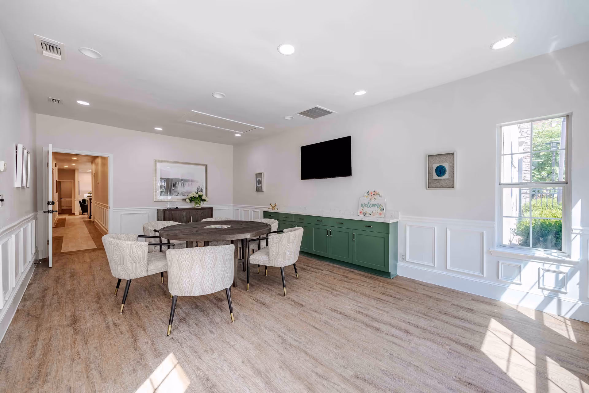Bright communal dining/activity room with a round table and upholstered chairs, wall-mounted TV, green cabinetry, and a large window.