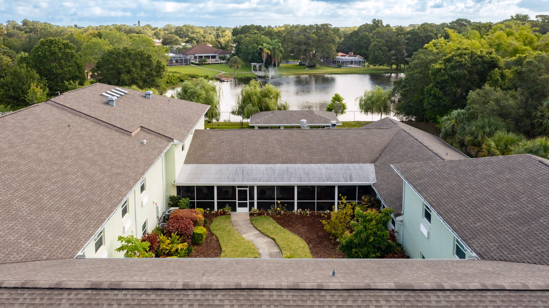 Aerial view of a senior living facility named Lake House showing a courtyard with a curved pathway, landscaped garden beds with shrubs and plants, surrounded by buildings with brown shingle roofs. In the background, there is a pond with a fountain, trees, and additional buildings.