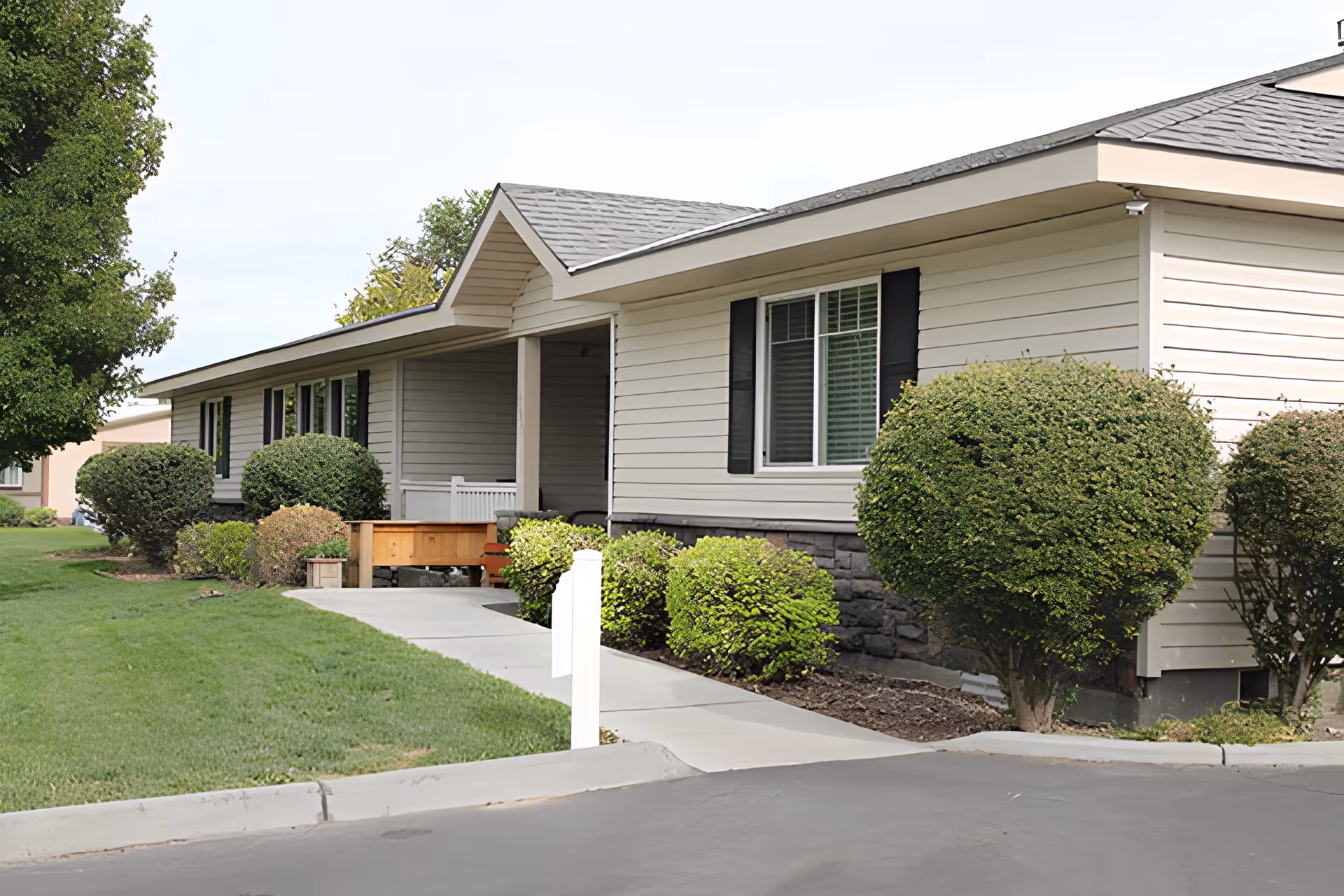 Exterior view of a single-story residential building with beige siding and a gray shingled roof. The building is surrounded by neatly trimmed bushes and a well-maintained lawn. A concrete walkway leads to a covered entrance with a small porch area.