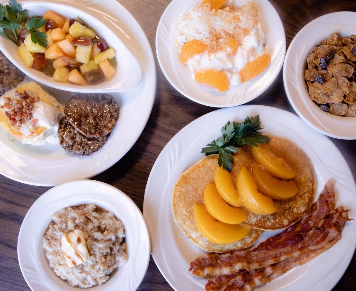 A breakfast meal consisting of pancakes topped with peach slices and garnished with parsley, crispy bacon strips, a bowl of oatmeal with a pat of butter, a plate with two sausage patties, a biscuit topped with gravy and bacon bits, a bowl of mixed fruit salad, a bowl of yogurt with mandarin orange slices and shredded coconut, and a bowl of raisin bran cereal.