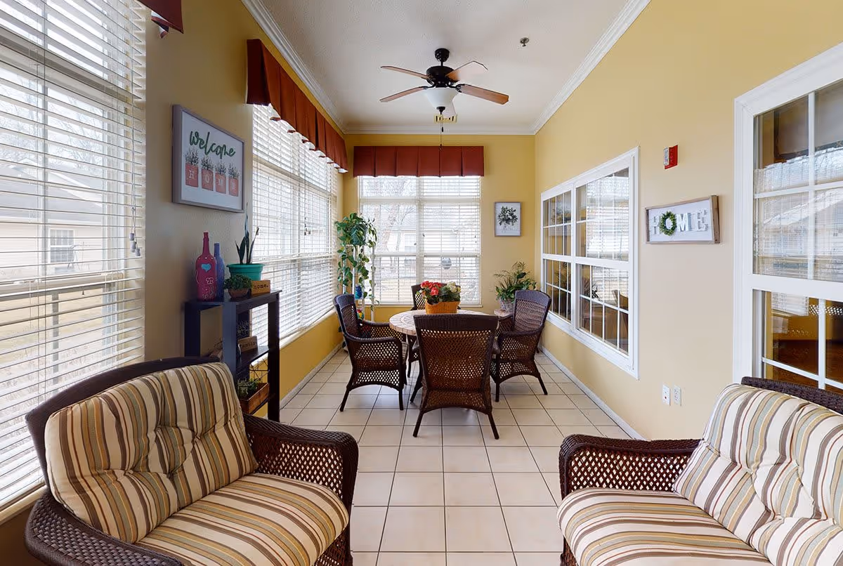 A bright sunroom with large windows covered by white blinds and red valances. The room features two cushioned wicker armchairs with striped upholstery, a round table with four wicker chairs, and several potted plants. The walls are painted yellow and decorated with framed signs, including one that says 'welcome home' and another that says 'HOME' with a wreath. The floor is tiled, and a ceiling fan is mounted above.