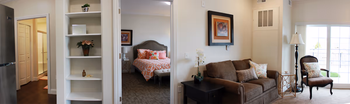 Interior view of a senior living facility showing a living room with a brown sofa, an armchair, a floor lamp, and a side table with a plant. To the left, there is a doorway leading to a bedroom with a bed dressed in orange and white patterned bedding and decorative pillows. There is also a white shelving unit with decorative items and a hallway with closed doors visible in the background.