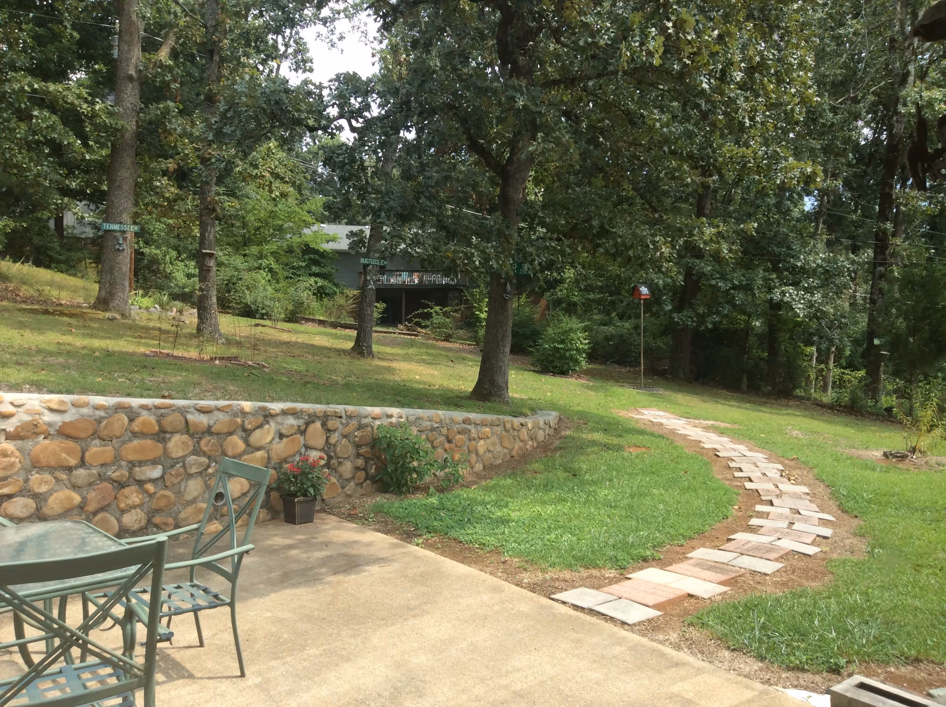 Outdoor patio area with a glass table and metal chairs on a concrete surface next to a stone retaining wall. A curved stone pathway leads through a grassy yard with trees and a birdhouse on a pole in the background.