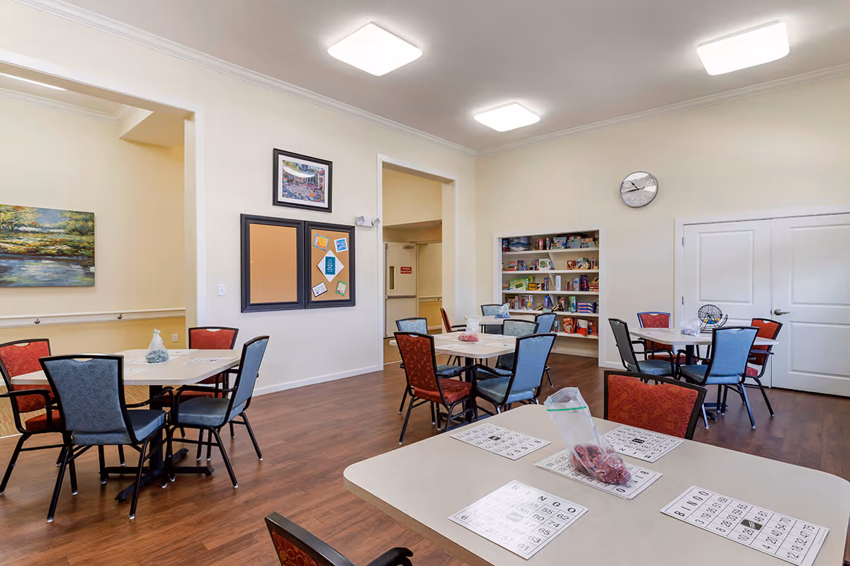 A well-lit common room with several tables and chairs arranged for group activities. The tables have bingo cards and a bag of bingo chips on them. The room features a bulletin board, a clock on the wall, and shelves stocked with board games and books. The floor is wooden, and the walls are painted a light color.