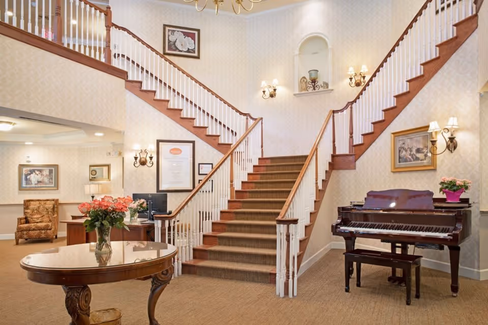 Interior view of a senior living facility lobby featuring a central carpeted staircase with wooden railings splitting into two directions at the top. A polished wooden table with a vase of pink flowers is in the foreground. To the right, there is a grand piano with a small bench and a potted plant on top. The walls are decorated with framed artwork and wall sconces with lampshades providing warm lighting. A comfortable armchair and a desk with a computer are visible in the background.