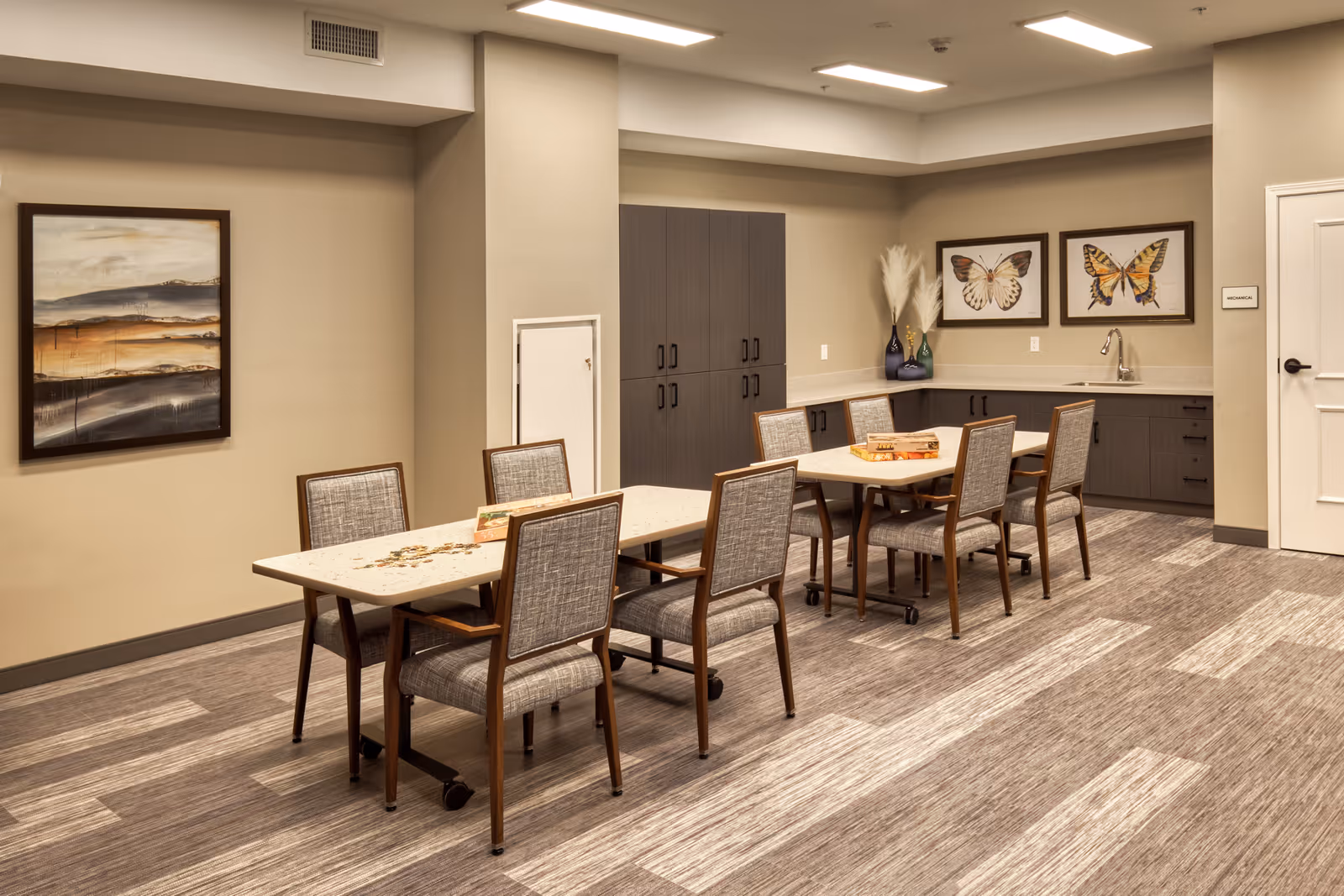 A well-lit room with two rectangular tables and eight cushioned chairs arranged around them. The room has beige walls, a patterned carpet, and dark wood cabinets along the back wall with a small sink. There are two framed butterfly pictures on the wall above the cabinets and a landscape painting on the adjacent wall. The door on the right side has a sign labeled 'MECHANICAL'.