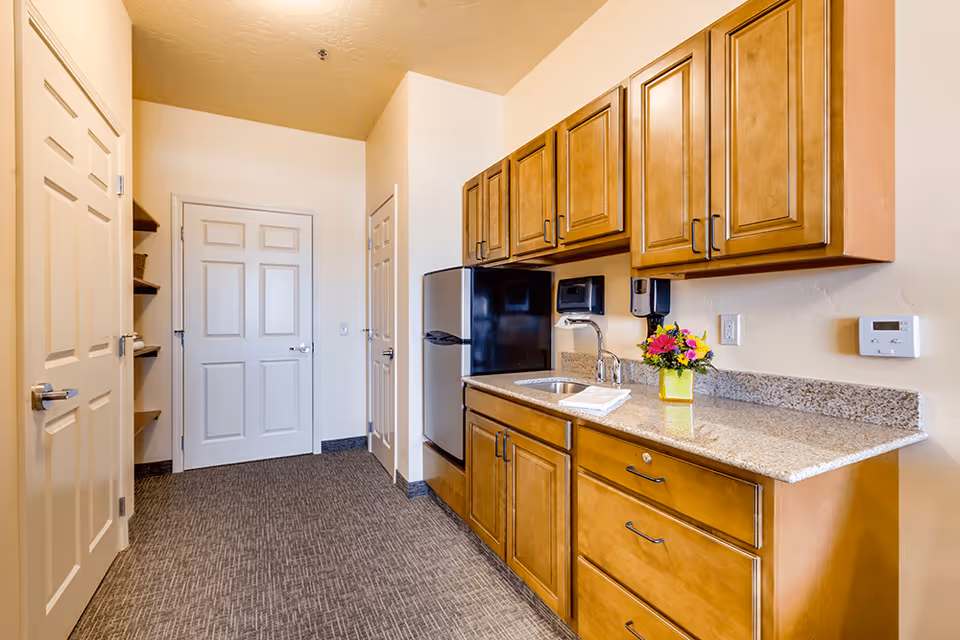 A compact kitchenette with wood cabinets, granite countertop, sink, mini refrigerator, and a small vase of flowers.