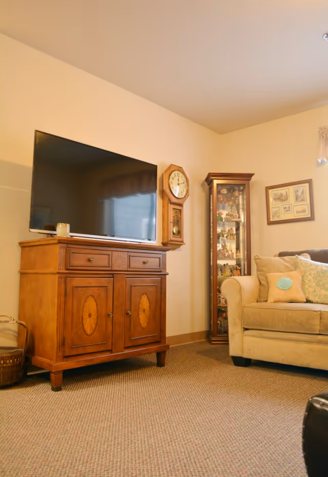 A cozy living room corner featuring a wooden cabinet with a flat-screen TV on top, a wall-mounted wooden clock, a glass display cabinet filled with collectibles, and a beige sofa with decorative pillows. The room has beige walls and carpeted flooring.