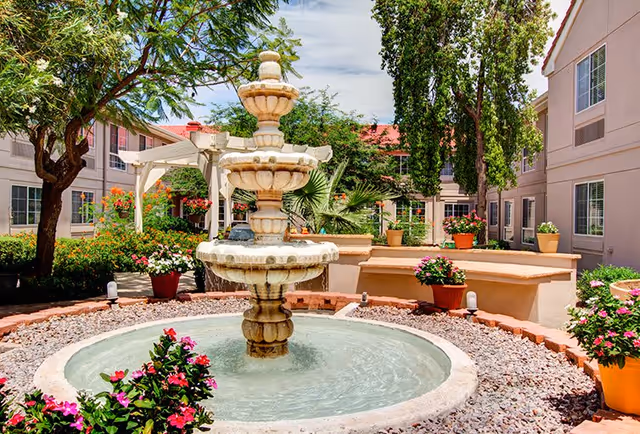 Outdoor courtyard area at The Gardens of Sun City featuring a three-tiered stone fountain surrounded by colorful potted flowers, trees, and shrubs, with beige buildings and windows in the background under a partly cloudy sky.