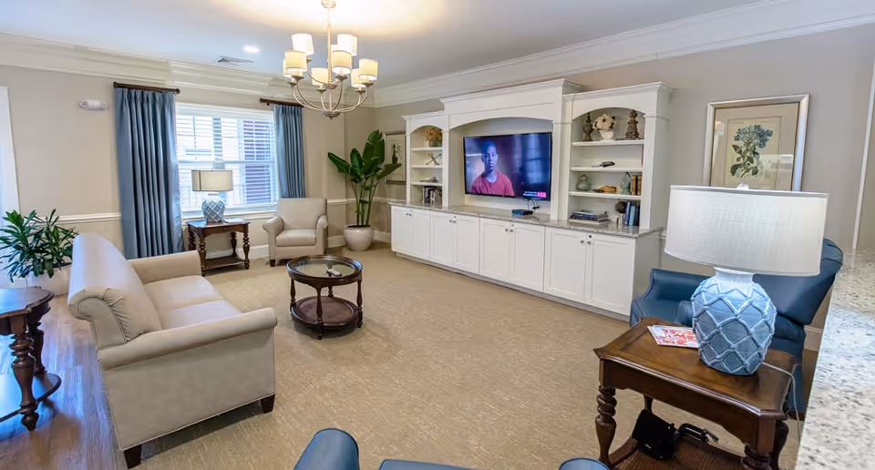 A cozy living room with beige walls and carpet, featuring a beige sofa, armchair, and two blue chairs. There is a round wooden coffee table in the center and side tables with lamps. A large built-in white entertainment center with shelves and cabinets holds a flat-screen TV displaying a person. Blue curtains frame a window, and there are decorative plants and framed artwork on the walls.