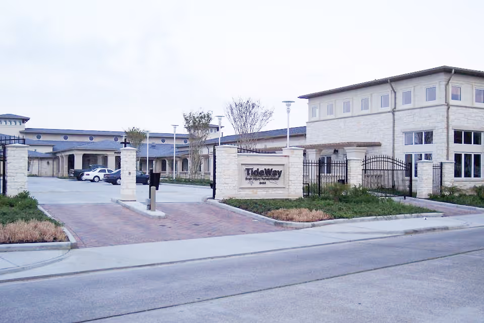 Exterior view of the Moody Neurorehabilitation Institute at TideWay showing a large building with stone facade, gated entrance, and a sign that reads 'TideWay Brain Injury Rehabilitation'. There are a few cars parked inside the gated area and some landscaping around the entrance.