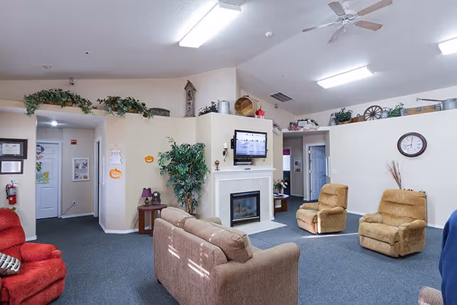 Communal living room with sofas and recliners arranged around a fireplace with a wall-mounted TV and decorative plants.