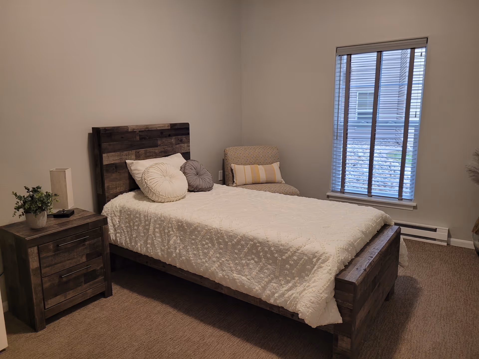 A bedroom with a wooden bed frame and headboard, a white textured bedspread, and two round decorative pillows. Next to the bed is a wooden nightstand with a small lamp and a potted plant. There is a patterned armchair with a striped lumbar pillow near a window with blinds, showing an exterior view of rocks and a building.