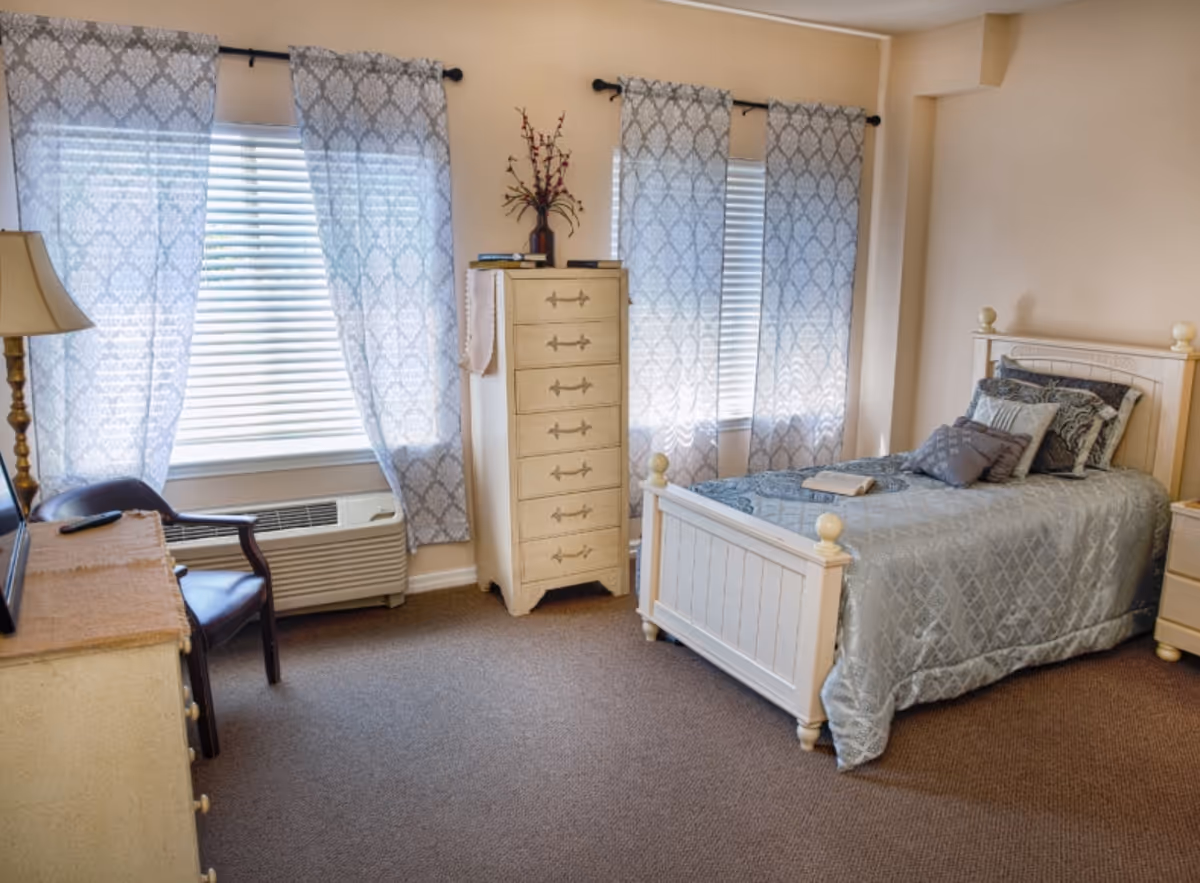 A cozy bedroom with a single bed covered in a light blue patterned bedspread and several pillows. The room has beige walls and carpeted floor. There are two windows with white blinds and light blue patterned curtains. A tall cream-colored dresser with decorative handles stands between the windows, topped with a vase of flowers and some books. A wooden chair and a small dresser with a lamp are positioned near one window.
