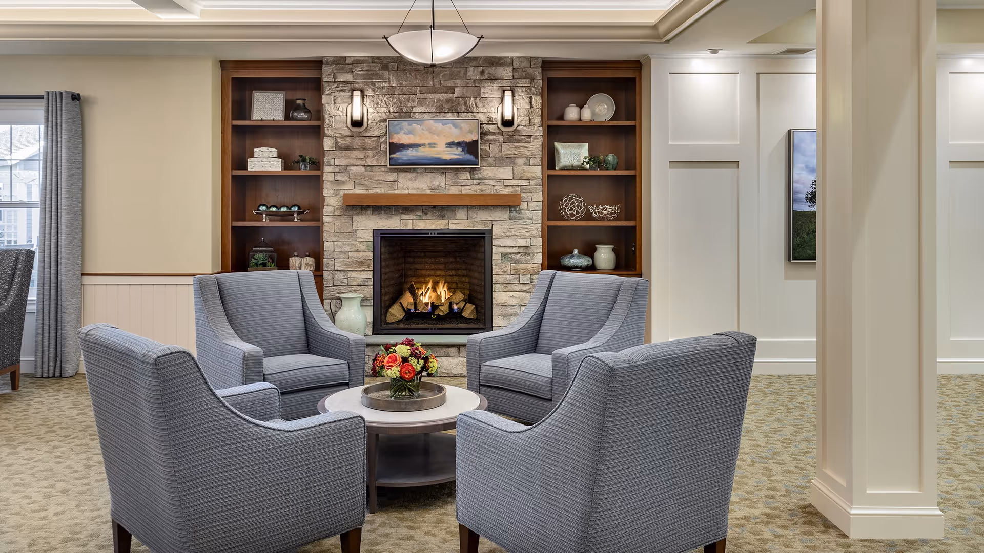 Cozy seating area with four gray armchairs arranged around a round coffee table in front of a stone fireplace with built-in shelves.