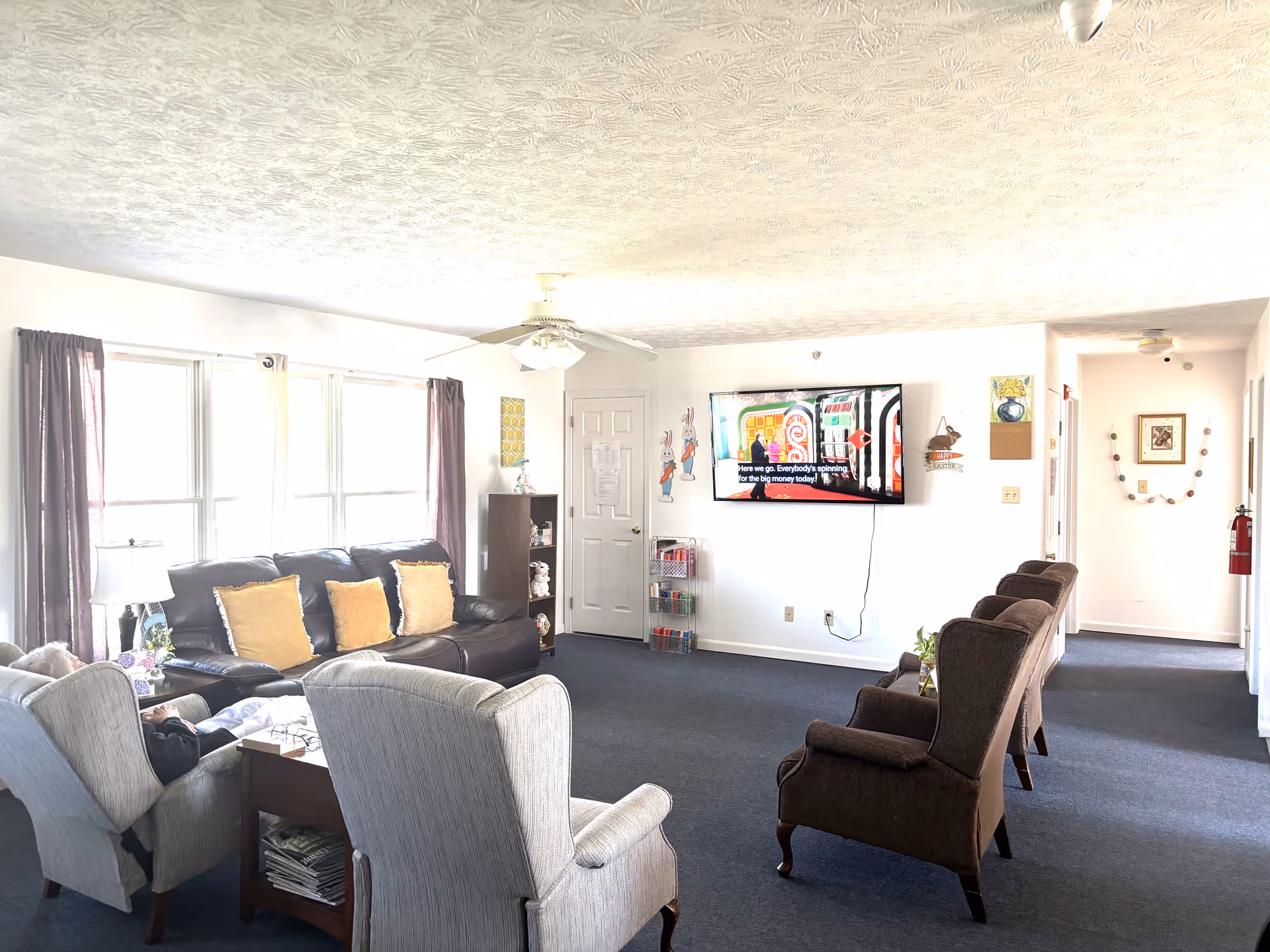 A bright living room in an assisted living facility with a large window covered by curtains on the left side. The room features a dark leather sofa with three yellow cushions, two light-colored armchairs, and three dark brown armchairs arranged facing a wall-mounted flat-screen TV. The TV is displaying a colorful game show with subtitles. The walls are decorated with small artworks and Easter-themed decorations. A ceiling fan is mounted on the textured ceiling, and a fire extinguisher is visible in the hallway on the right.