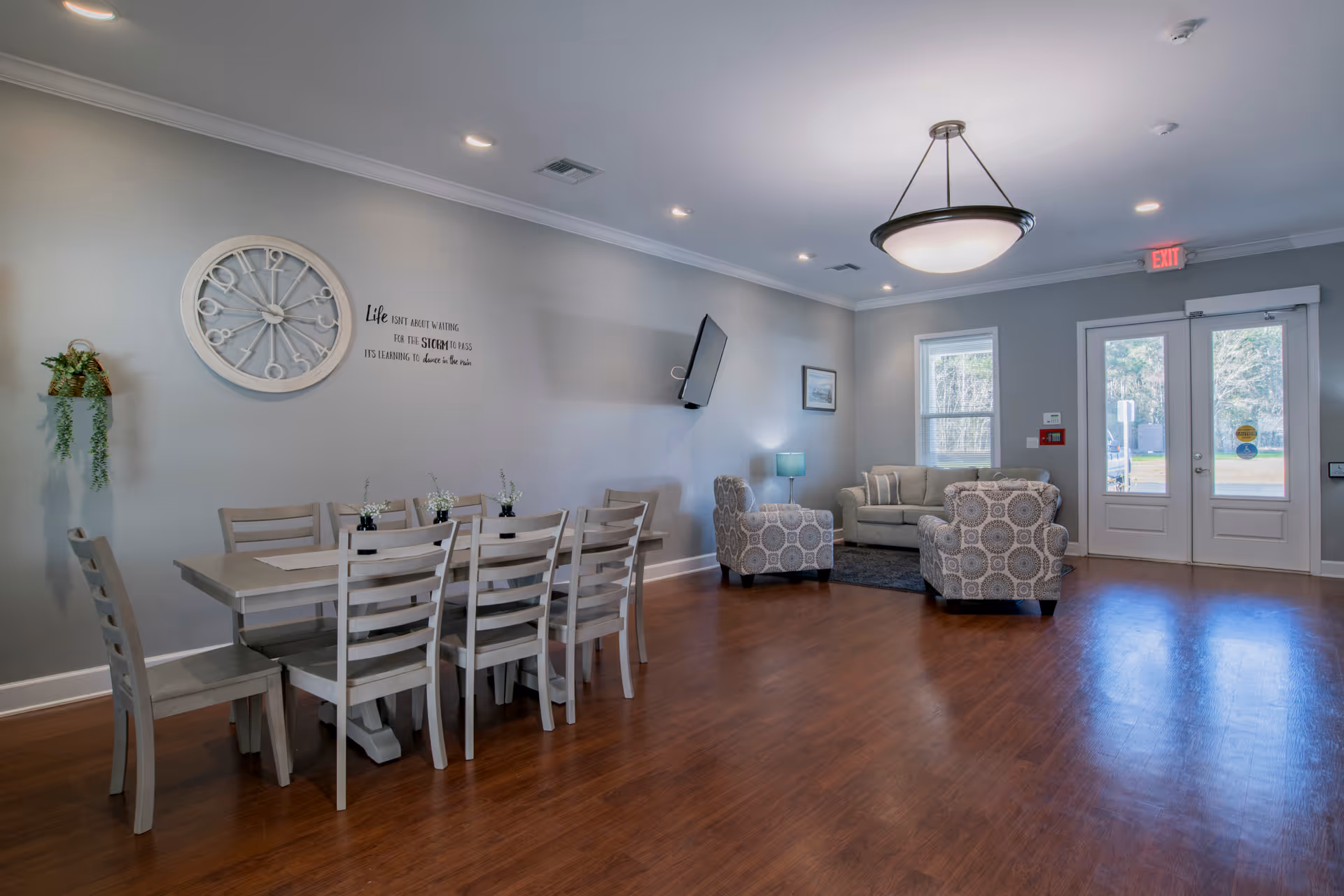 A spacious and well-lit common area with a long dining table and eight chairs on the left side, decorated with small plants and a large wall clock above. On the right side, there is a seating area with a sofa, two patterned armchairs, a small lamp on a side table, and a wall-mounted TV. The room has wooden flooring, light gray walls, recessed ceiling lights, a hanging ceiling light fixture, and double glass doors leading outside.