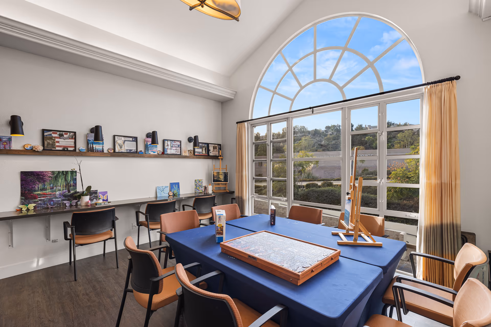 A bright activity room with a large arched window letting in natural light. The room has a table covered with a blue tablecloth, surrounded by chairs, with a puzzle and an easel on top. Along one wall, there is a long shelf with various framed pictures and small decorative items, and chairs are placed underneath the shelf. Beige curtains frame the window, and the floor is wood.