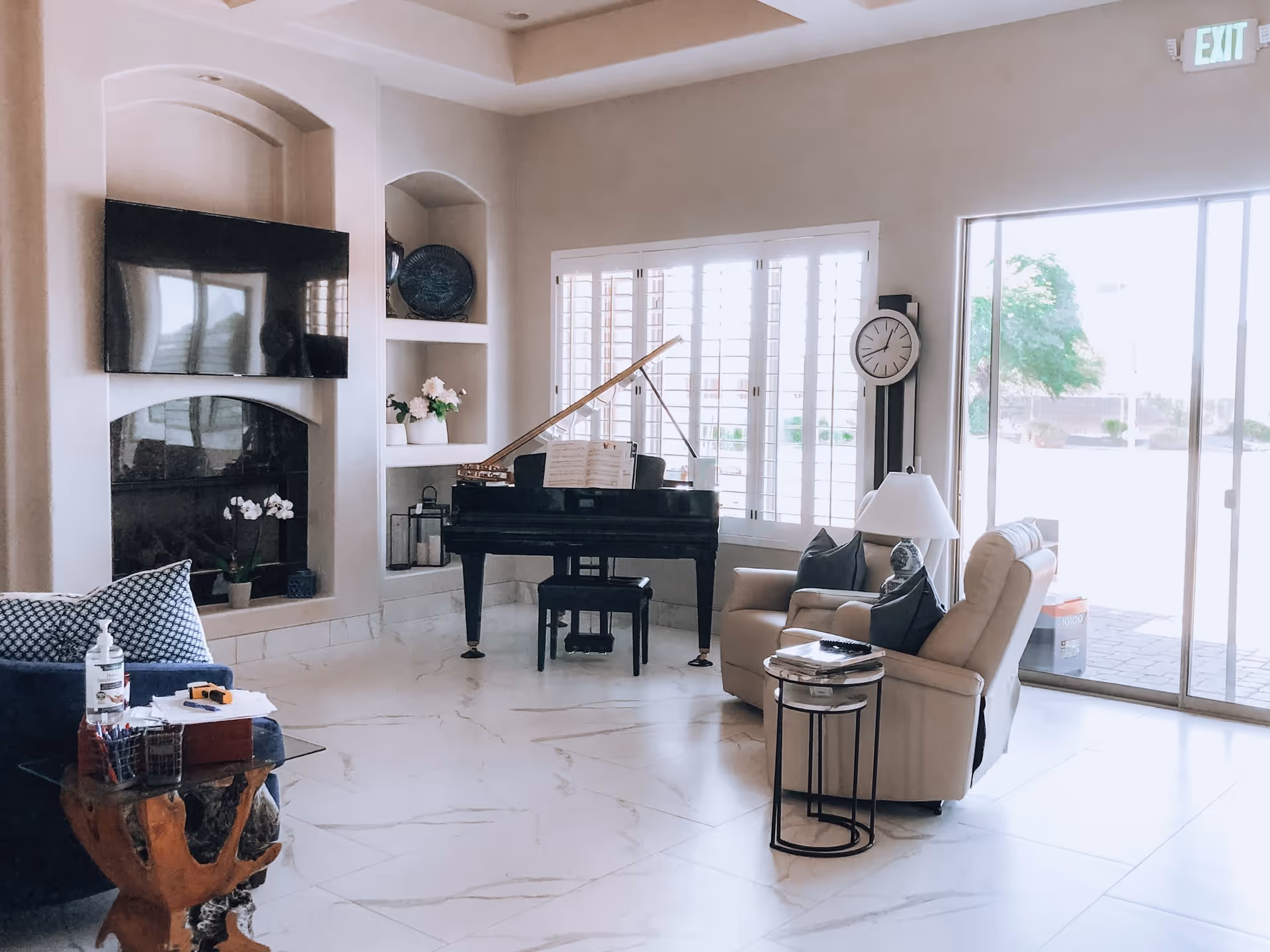 Bright living room with a grand piano by shuttered windows, a wall-mounted TV above a fireplace, reclining chairs, and a sliding glass door.