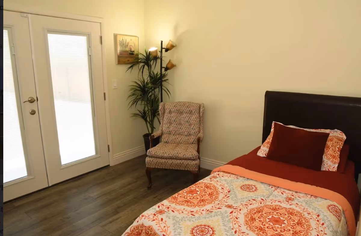 A bedroom with a single bed featuring a colorful patterned bedspread and pillows. Next to the bed is a patterned armchair, a tall floor lamp with three lights, and a potted plant. There is a set of double glass doors with blinds on the left side of the room, and a framed picture hanging on the wall above the chair.