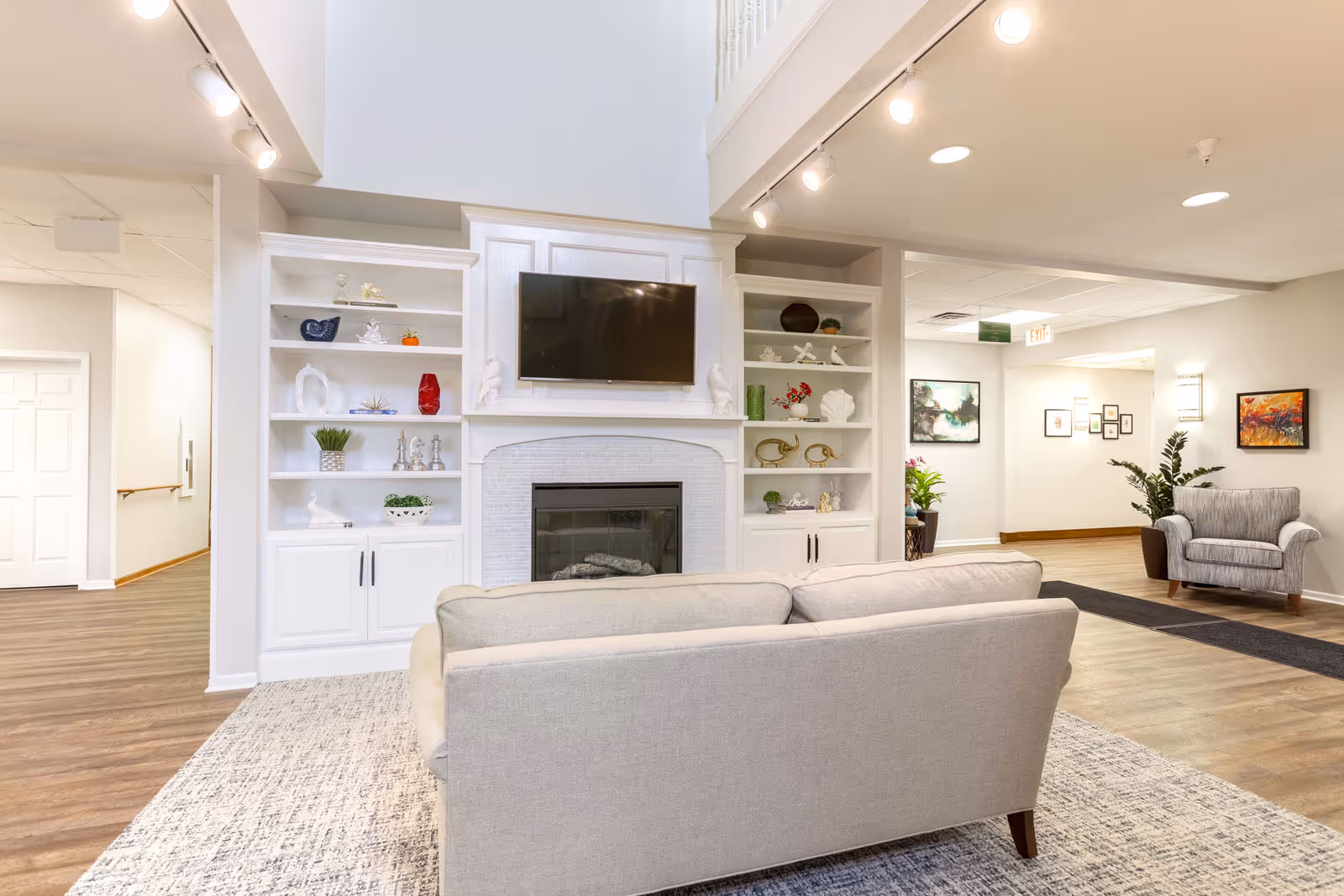 A bright and spacious living room area in a senior living facility with a beige sofa facing a wall-mounted flat-screen TV above a white fireplace. The fireplace is flanked by built-in white shelves decorated with various ornaments and plants. The room has wooden flooring, a patterned area rug, and additional seating including a gray armchair. The walls are adorned with framed artwork and there is a hallway visible in the background.