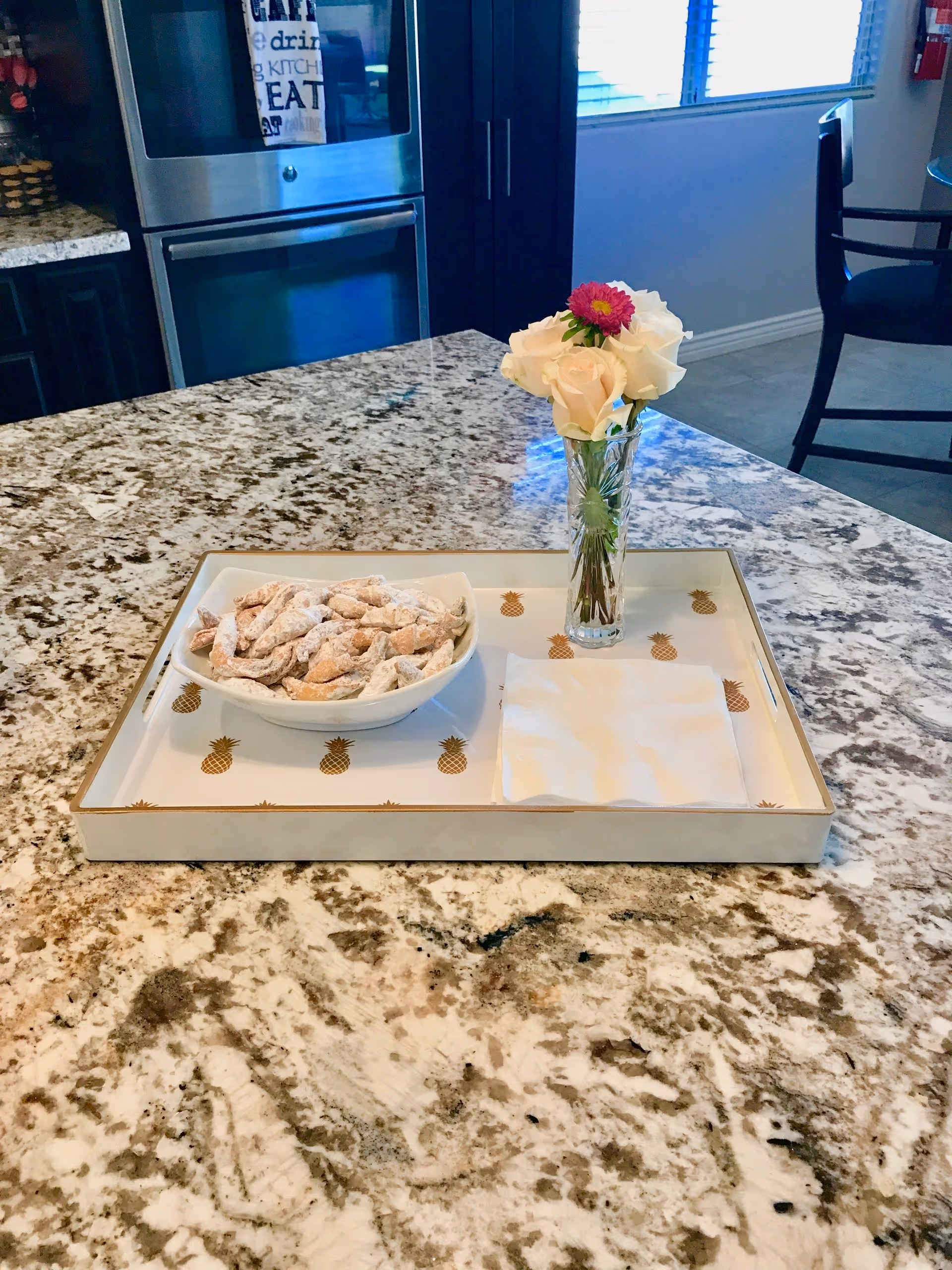 Granite kitchen island with a decorative tray holding a bowl of powdered pastries and a small vase of flowers, with an oven and seating visible in the background.
