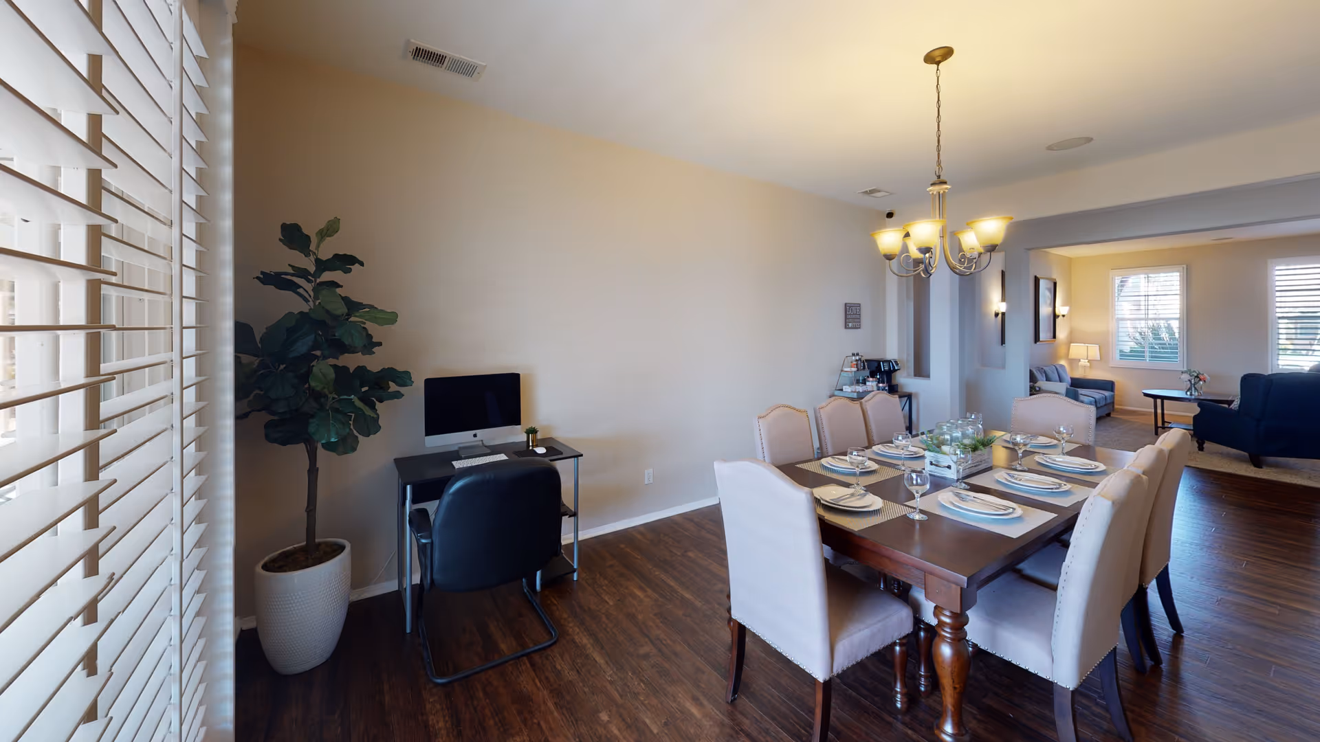 Dining room with a set wooden table and chandelier, an adjacent living area, and a small computer desk and potted plant by window blinds.
