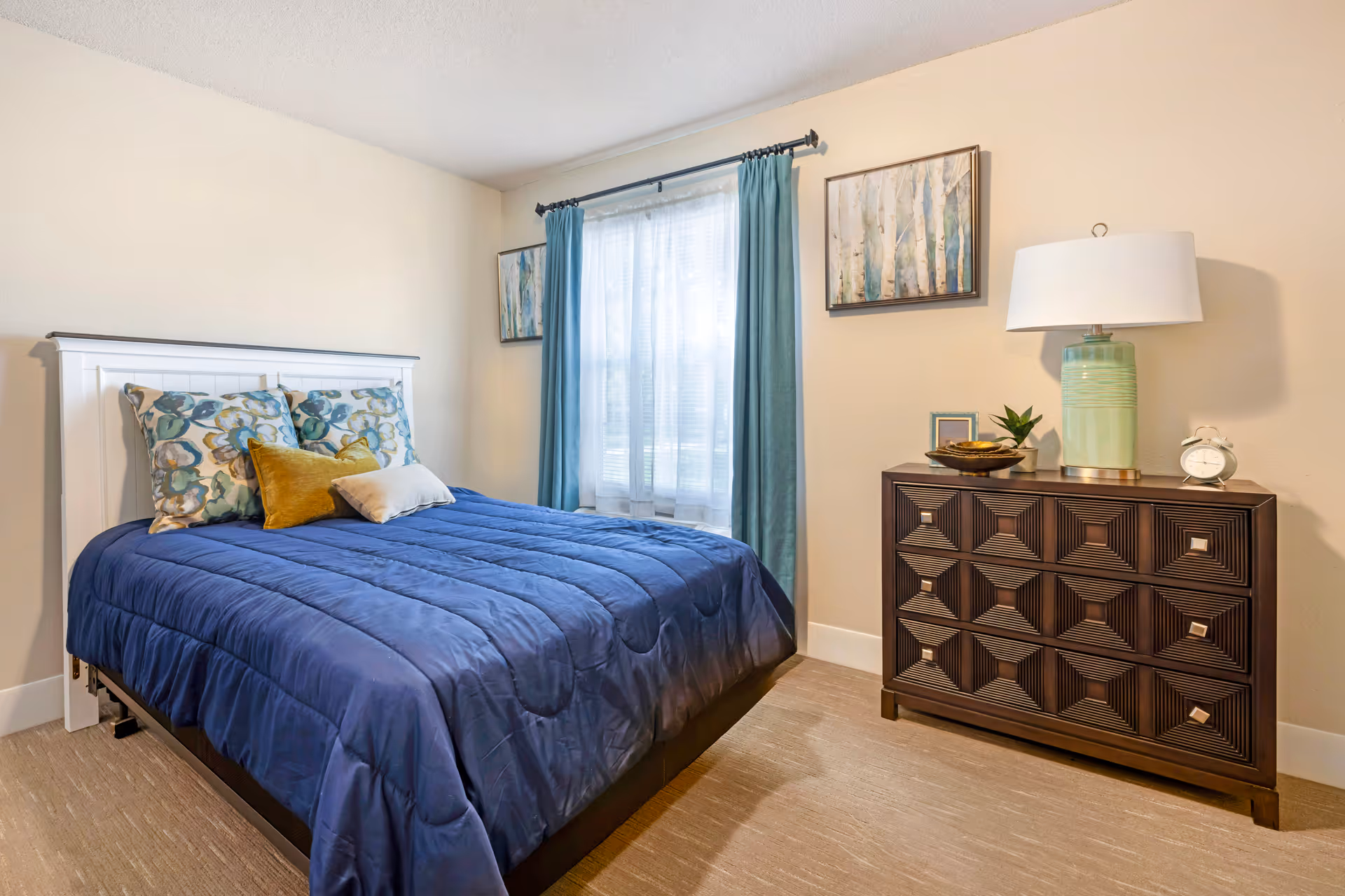 A bedroom with a bed covered in a blue comforter and decorative pillows. There is a window with sheer white curtains and blue drapes. On the right side, there is a dark wooden dresser with a green lamp, a small plant, a picture frame, and an alarm clock. Two framed paintings hang on the wall above the dresser and beside the window.