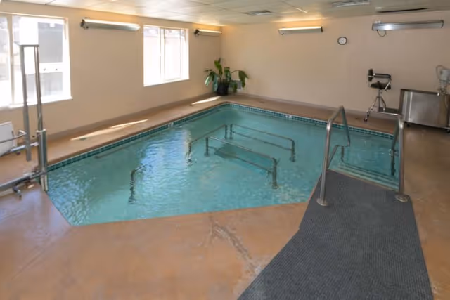 Indoor rehabilitation pool with handrails, a lift, and windows in a well-lit therapy room.