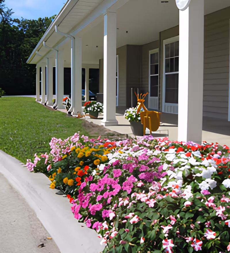 A colorful flower bed with pink, white, red, yellow, and purple flowers in front of a covered porch with white columns at Pleasant View Assisted Living. The porch has potted plants and a decorative orange metal deer sculpture.