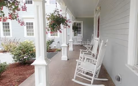 A covered porch area with white rocking chairs lined up along the wall of a light gray building. There are white columns supporting the porch roof and hanging flower baskets. The porch overlooks a landscaped area with bushes and flowers.