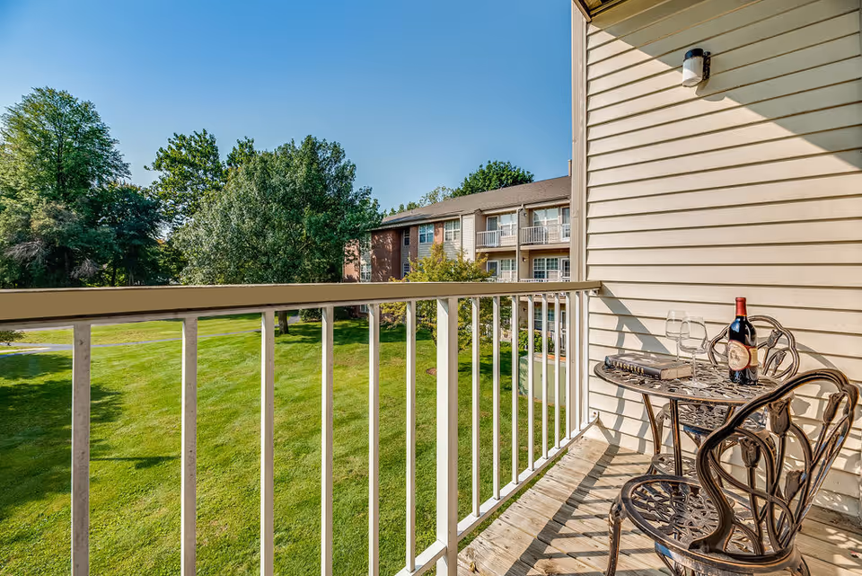 A balcony with a small ornate metal table and two matching chairs. On the table, there is a bottle of red wine, two empty wine glasses, and a book. The balcony overlooks a green lawn with trees and another residential building in the background under a clear blue sky.