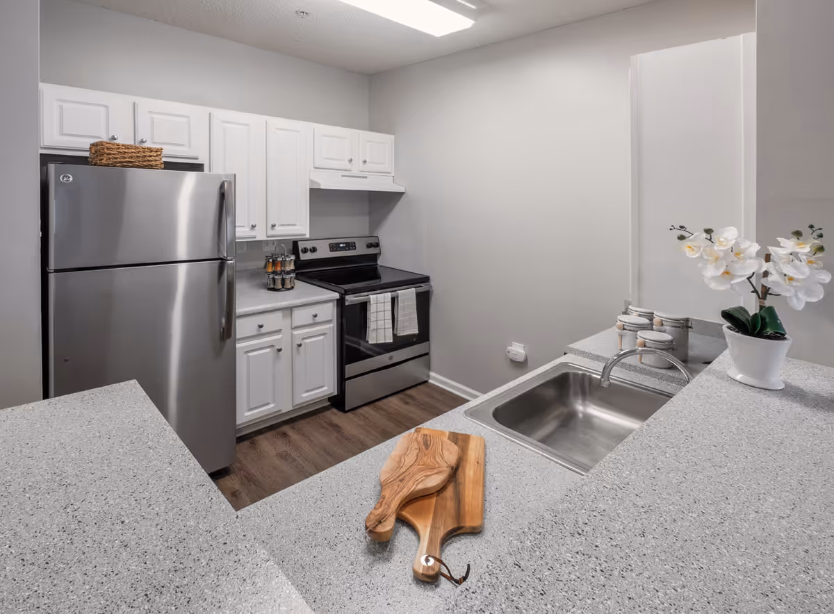 Modern kitchen with stainless steel refrigerator and stove, white cabinets, gray countertops, a sink with a faucet, a wooden cutting board, and a white orchid plant in a pot.