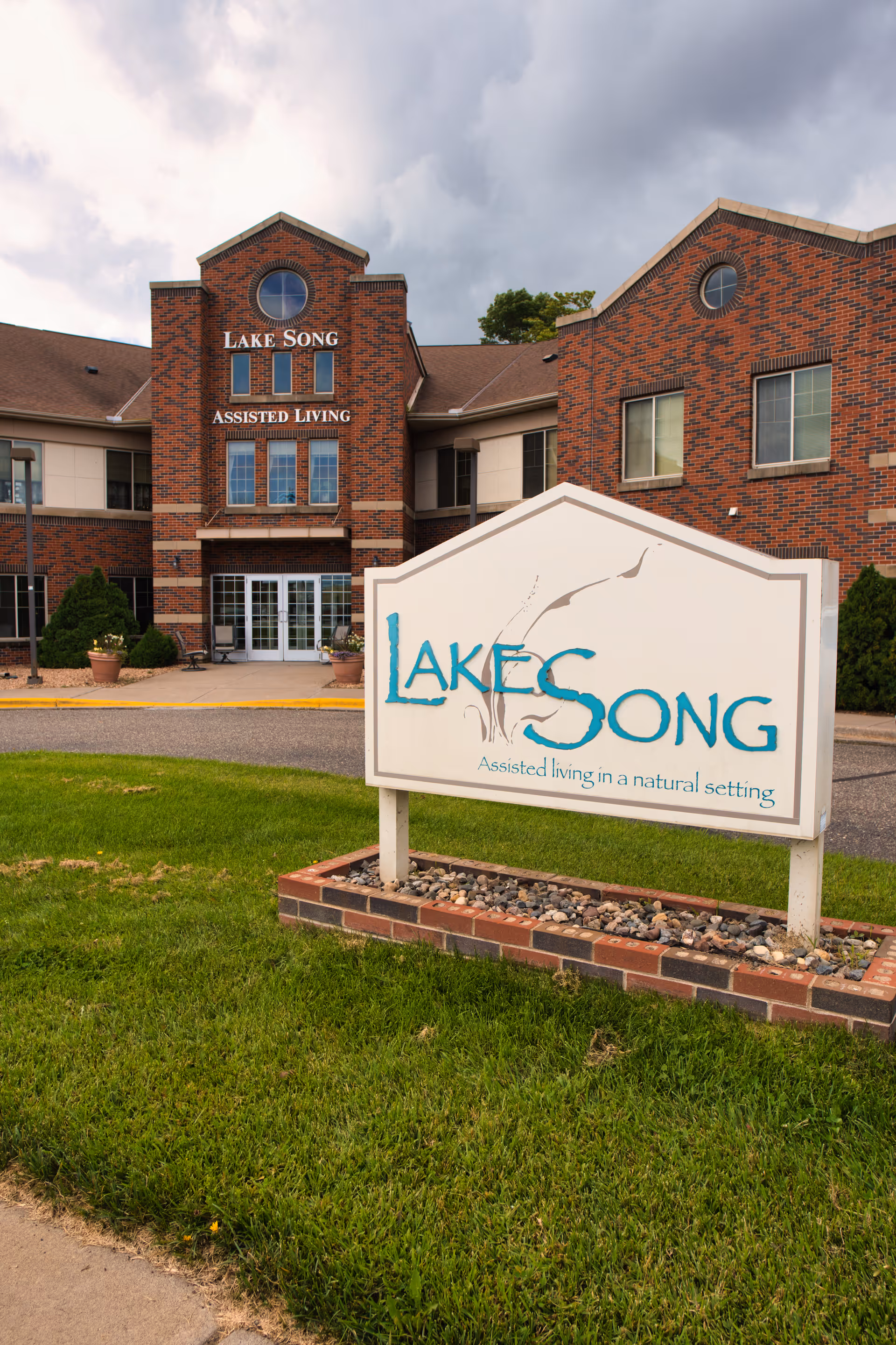 Front entrance of the Lake Song Assisted Living building with a large freestanding sign on the lawn in the foreground.