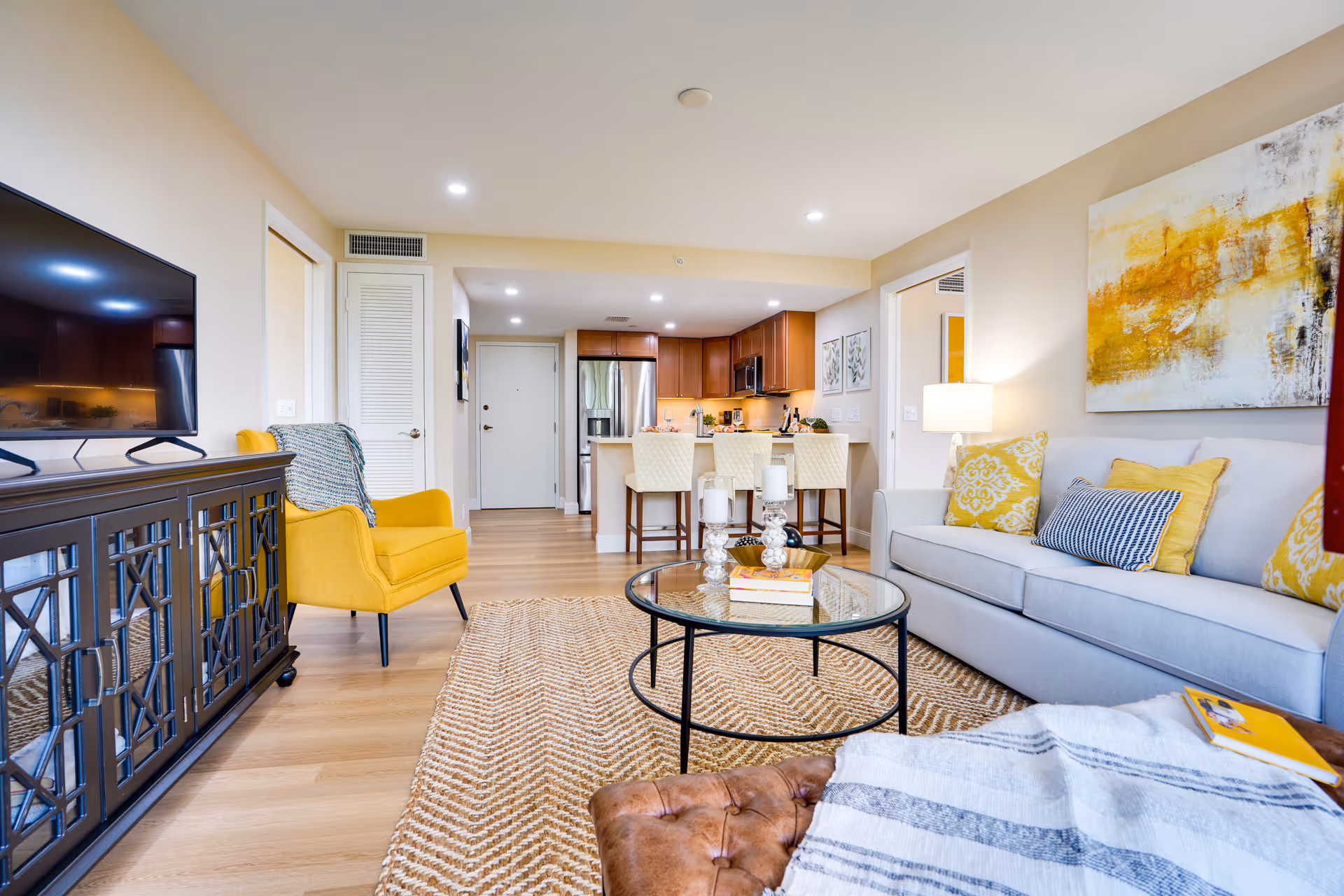 A bright and modern living room with a light gray sofa adorned with yellow and blue patterned pillows, a round glass coffee table with candles and books, a yellow armchair, a large flat-screen TV on a dark wooden cabinet, and a view into a kitchen area with wooden cabinets and a breakfast bar with three cream-colored chairs.