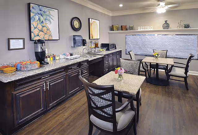 A cozy dining area with two square tables, each with a small vase of flowers. The room features dark wooden chairs with patterned cushions and a long granite countertop with cabinets underneath. On the countertop, there are various items including a coffee dispenser, cups, and a microwave. The walls are decorated with framed artwork and a clock, and a shelf above the window holds decorative items and letters spelling 'BISTRO'. The floor is wooden, and the window is covered with a textured blind.