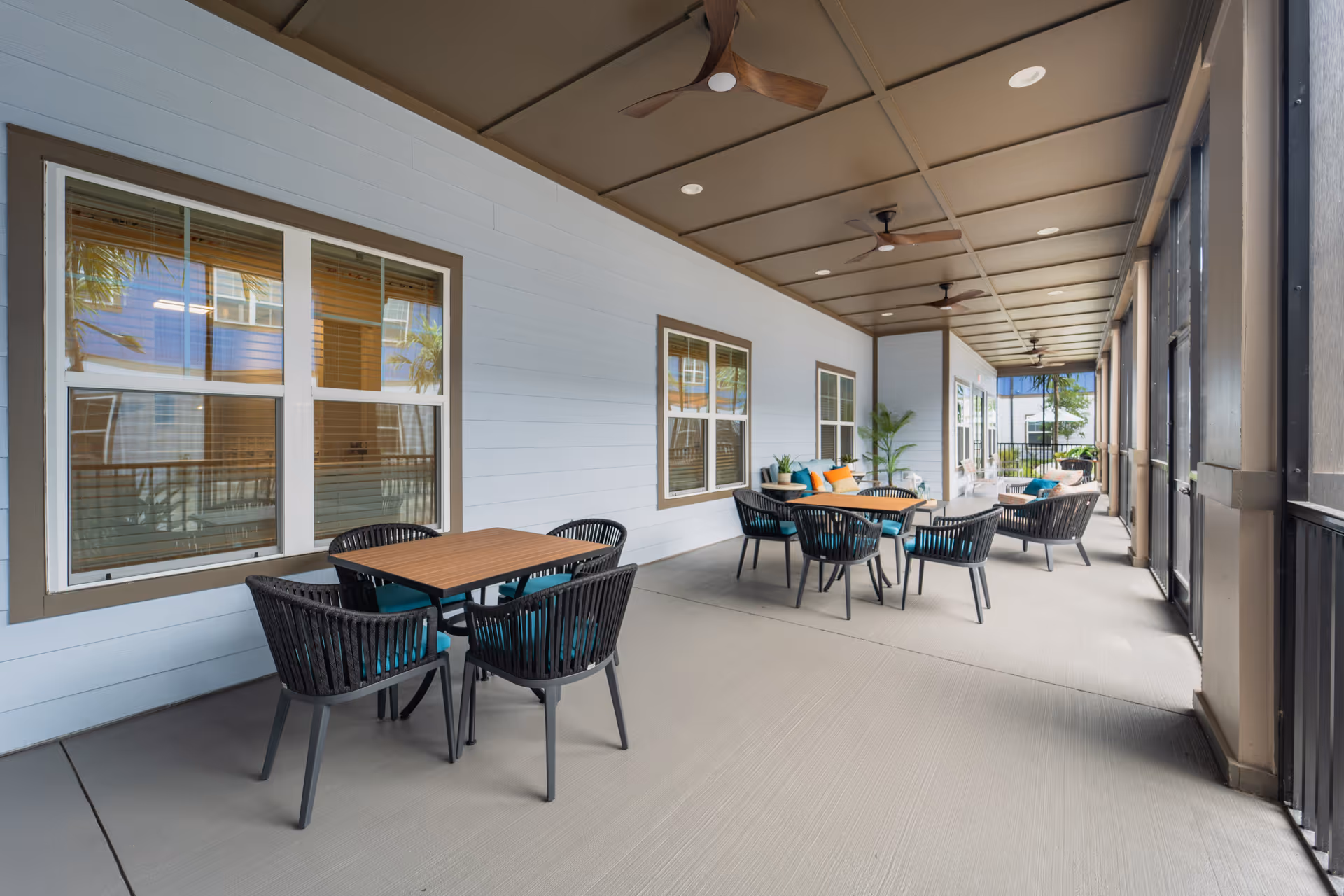 A covered outdoor patio area with several tables and chairs arranged for seating. The space has ceiling fans, potted plants, and large windows along one wall. The patio is enclosed with screened panels on the right side.