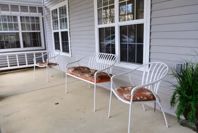 A covered outdoor patio area with three white metal chairs and one white metal bench, all with floral-patterned cushions. The patio is adjacent to a building with light gray siding and multiple large windows. There is a green potted plant on the right side of the image.