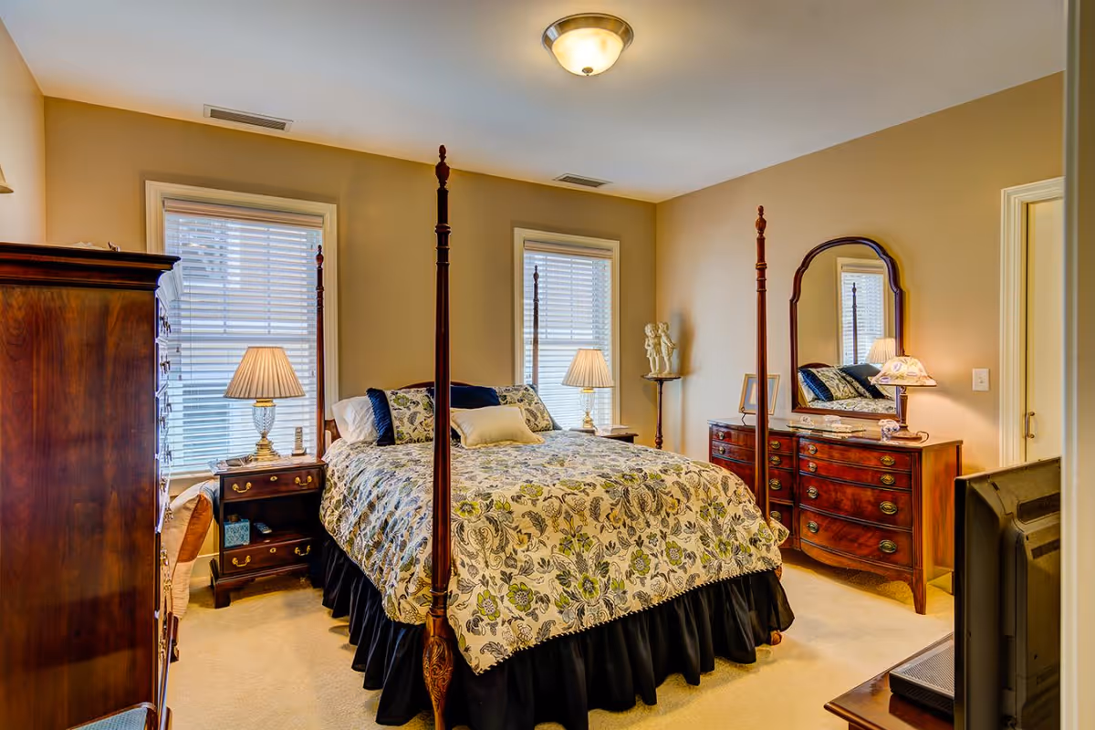 A cozy bedroom with a four-poster bed covered in a floral patterned bedspread. The room has two windows with white blinds, two bedside tables with lamps, a wooden dresser with a mirror, and a TV on a stand. The walls are painted beige and the carpet is light-colored.