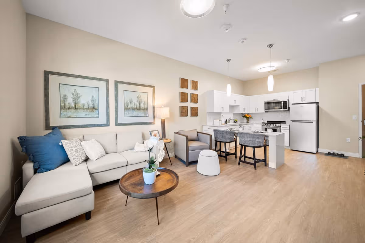 A bright and modern living room and kitchen area featuring a beige sectional sofa with blue and white pillows, a round wooden coffee table with a potted plant, a gray armchair, and a white ottoman. The kitchen has white cabinets, stainless steel appliances including a refrigerator, microwave, and stove, and a small island with two bar stools. The walls are decorated with framed artwork and wooden wall hangings, and the floor is light wood.