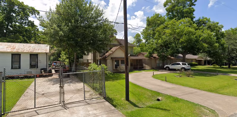 Front exterior of a residential house with a gated driveway, curved concrete drive, parked vehicles, and large trees.
