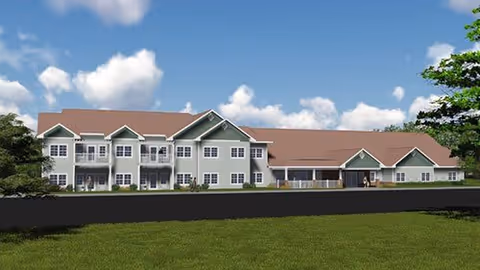 Exterior view of a two-story senior living facility building with a green lawn in the foreground, multiple windows, balconies, and a brown roof under a partly cloudy blue sky.