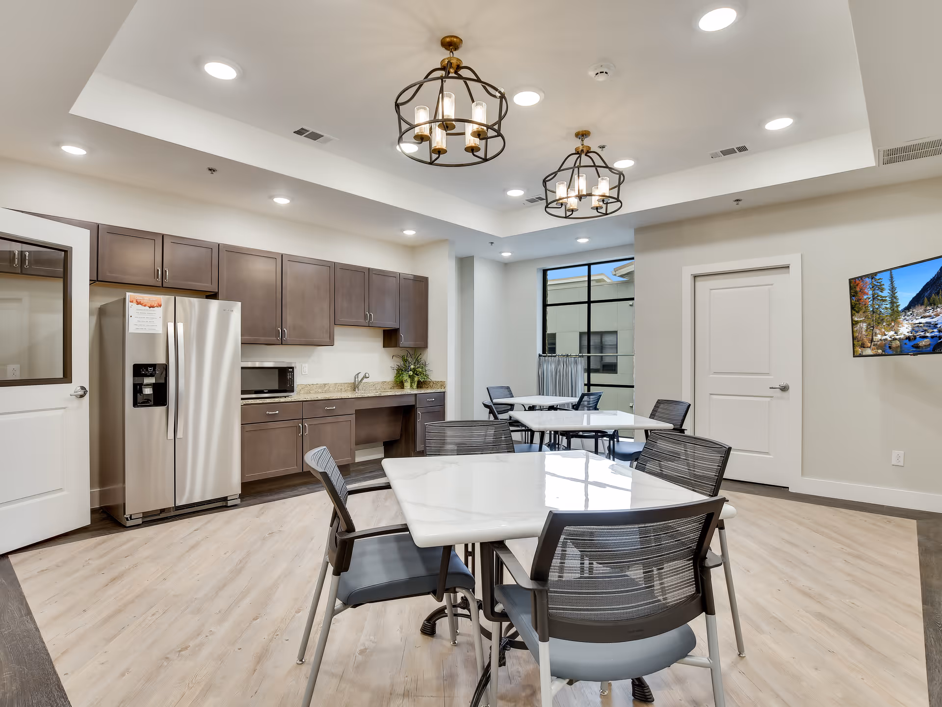 A modern dining area with white square tables and black mesh chairs. The room features a kitchen area with brown cabinets, a stainless steel refrigerator, a microwave, and a granite countertop. Two decorative ceiling light fixtures hang above, and a large window lets in natural light. A flat-screen TV is mounted on the wall showing a scenic nature image.