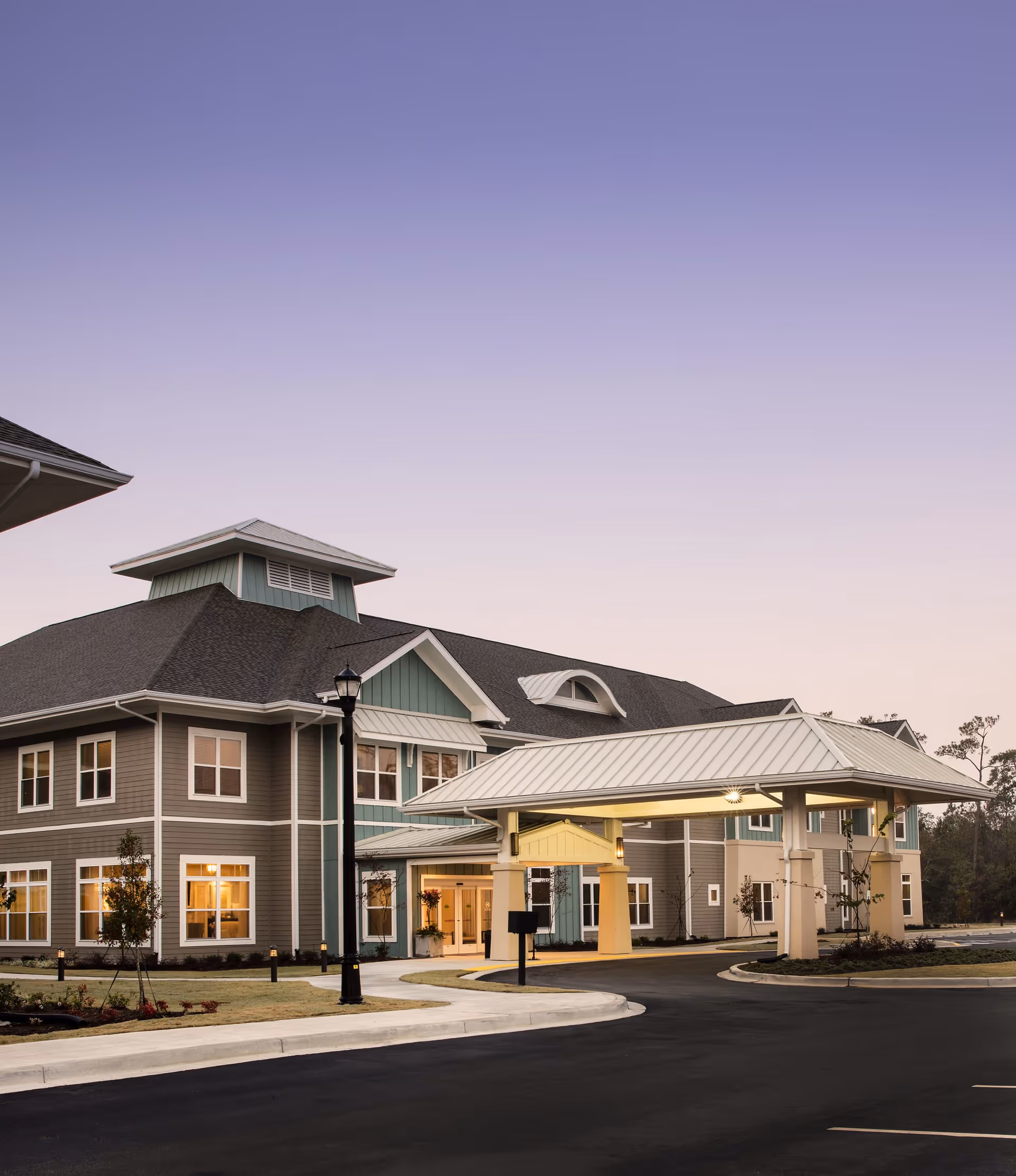 Exterior view of a senior living facility building at dusk with a covered entrance driveway, multiple windows, and a well-maintained surrounding landscape.